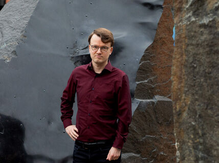 Person in a maroon shirt standing next to a large textured rock with one hand on hip.