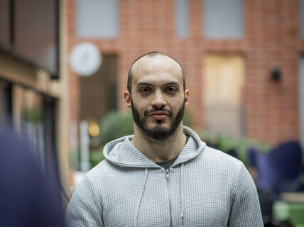 A person wearing a light grey hoodie stands indoors with a brick wall and green plants in the background.