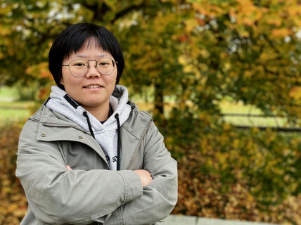 Person standing outdoors in autumn, wearing a grey hoodie and green jacket. Trees in the background with orange leaves.