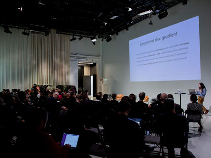 Audience in a large room facing a screen with a person speaking at the front of the room