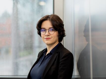 A person with short dark hair wearing a black blazer and navy shirt stands next to a window with a blurred background.