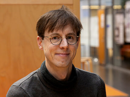 A person wearing a dark grey turtleneck sweater is indoors. The background includes wooden panels and glass walls with posters.