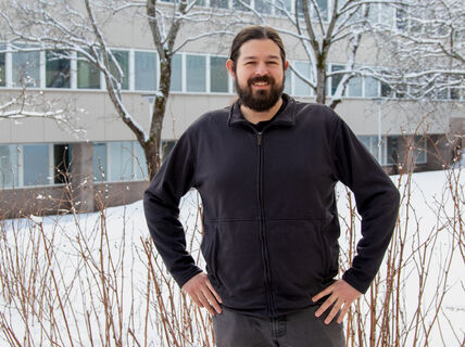 Person in dark jacket stands in snowy yard before office building with bare, snow-covered trees