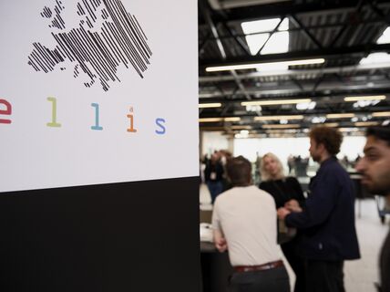 Sign with striped Europe map and colourful “ellis” logo beside people talking in a bright hall