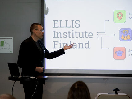 Person in dark suit giving a presentation about ELLIS Institute Finland on a bright screen
