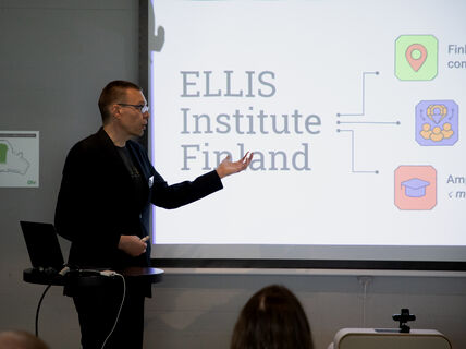 Person in dark suit giving a presentation about ELLIS Institute Finland on a bright screen