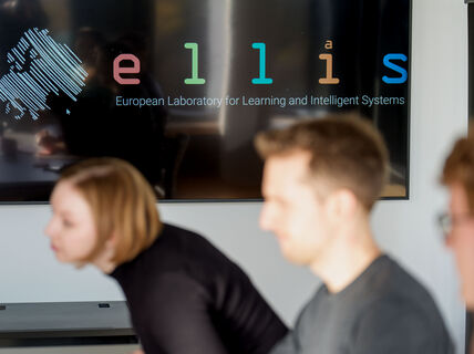 Three people sitting in a meeting room, ELLIS logo is displayed on the screen.