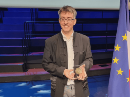 Man standing in auditorium holding medal, next to EU and French flags