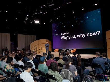 Seated audience facing a person standing on a stage in front of a large screen