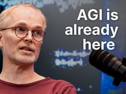Middle‑aged man in glasses speaks into a black studio microphone, text reads “AGI is already here”.