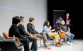 Five people seated in a semicircle of chairs in front of a screen. One person standing in front of a banner to the side.