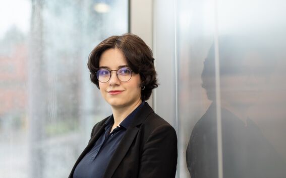 A person with short dark hair wearing a black blazer and navy shirt stands next to a window with a blurred background.