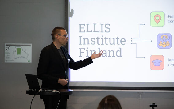 Person in dark suit giving a presentation about ELLIS Institute Finland on a bright screen
