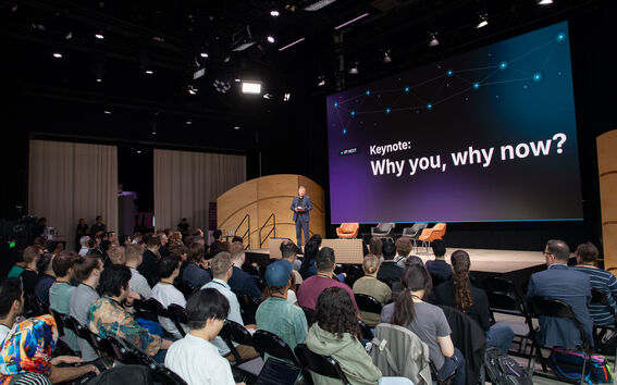 Seated audience facing a person standing on a stage in front of a large screen