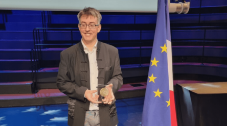 Man standing in auditorium holding medal, next to EU and French flags