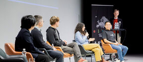 Five people seated in a semicircle of chairs in front of a screen. One person standing in front of a banner to the side.