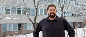 Person in dark jacket stands in snowy yard before office building with bare, snow-covered trees