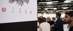 Sign with striped Europe map and colourful “ellis” logo beside people talking in a bright hall