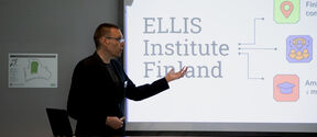 Person in dark suit giving a presentation about ELLIS Institute Finland on a bright screen