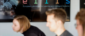 Three people sitting in a meeting room, ELLIS logo is displayed on the screen.
