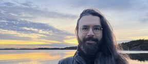 Person with long hair and beard standing against a sea background