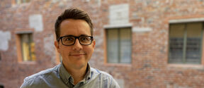 Man wearing glasses and button-down shirt against a brick building background