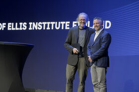 Two men smiling men standing in front of a large screeen 