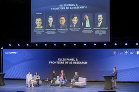 Five people sitting on sofas on a stage, with their photos projected overhead. One person standing on the right next to a podium