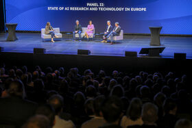 Five people sit on sofas on a stage with audience visible in foreground. Text in background reads PANEL II: STATE OF AI RESEARCH AND TECHNOLOGY IN EUROPE