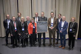 Group of people standing in front of curtains. Samuel Kaski in center holds a small square blackboard with the word LAUNCHED written on it
