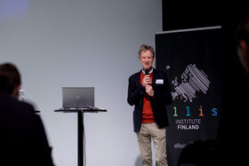 Man with a microphone, standing next to a laptop and a banner