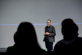 Man speaking in front of a screen with two people in shadow in the foreground