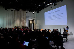Audience in a large room facing a screen with a person speaking at the front of the room