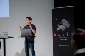 Man speaking at podium with raised hands. A laptop is in the podium and a banner is to the side
