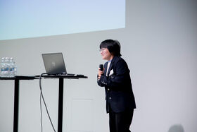 Woman holds a microphone while standing in front of a podium with a laptop