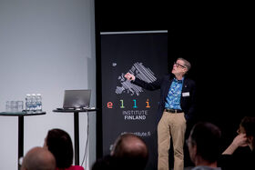 Man gestures towards a screen, with laptop on podium nearby