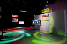 Dark room with red, turquoise and green light rings on the floor. Printed signs and screens are around the room.