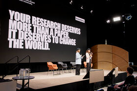 Two people speaking into microphones on a stage in front of a large screen