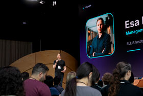 Man speaking into a microphone on a stage, standing next to a screen. Audience members are visible in the foreground.
