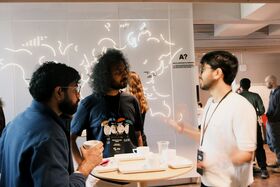 Three people standing around a table talking, with a white wall with world map in background