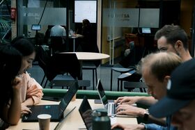 People working on laptops; tables, chairs and other rooms visible in background.