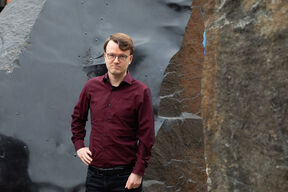 Person in a maroon shirt standing next to a large textured rock with one hand on hip.