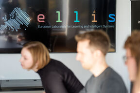 Three people sitting in a meeting room, ELLIS logo is displayed on the screen.