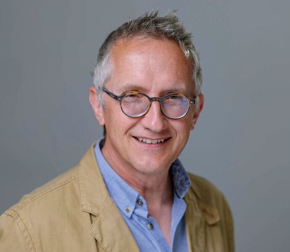 A person with grey hair wearing a beige jacket and light blue shirt