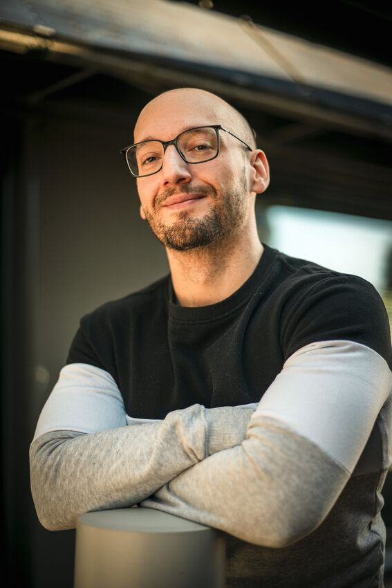 Person with crossed arms, wearing black and grey jumper outdoors by a building