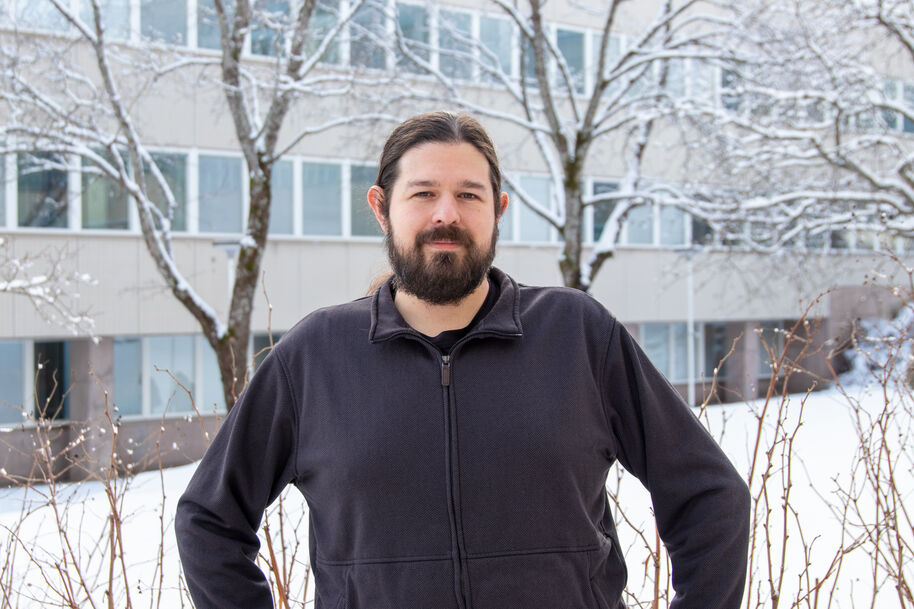 Person in dark jacket stands in snowy yard before office building with bare, snow-covered trees
